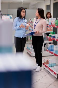 Pharmacy assistant and customer examining medicaments, looking at supplements box from shelves. Asian people talking about healthcare products and medicinal supplies, pharmaceutical drugs.