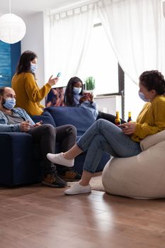 Mixed racial people sitting together in living room browsing on phones wearing protection masks, spending free time respecting social distance. Diverse people enjoying party during global pandemic