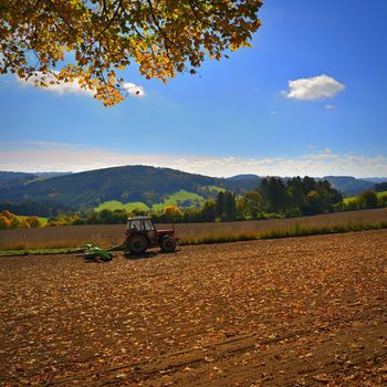 Tractor on the field. Beautiful autumn nature with landscape in the Czech Republic. Colorful trees with blue sky and sun. Background for autumn and agriculture.