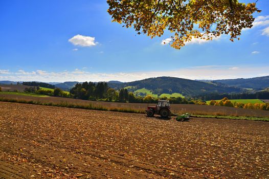 Tractor on the field. Beautiful autumn nature with landscape in the Czech Republic. Colorful trees with blue sky and sun. Background for autumn and agriculture.