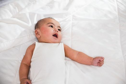 Asian cute little baby on bed under soft blanket, baby wearing bodysuit lying on white beedsheets in bedroom, smiling infant child at home