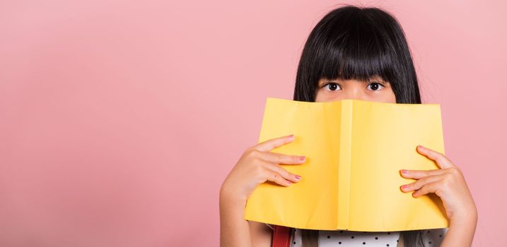 Asian little kid 10 years old holding and reading yellow book near eyes at studio shot isolated on pink background, Happy child girl read dictionary paper books, lifestyle lovely educated cheerful