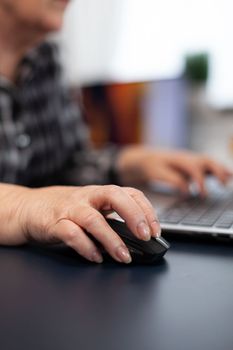 Close up of old entrepreneur lady using mouse in home office. Elderly woman in home living room using moder technoloy laptop for communication sitting at desk indoors.