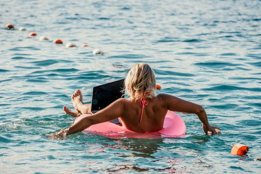 Woman works on laptop in sea. Freelancer, young blond woman in sunglases floating on an inflatable big pink donut with a laptop in the sea at sunset. Freelance, travel and holidays concept