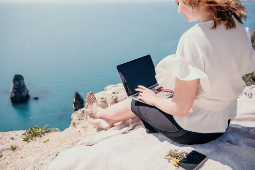 Successful business woman in yellow hat working on laptop by the sea. Pretty lady typing on computer at summer day outdoors. Freelance, travel and holidays concept.