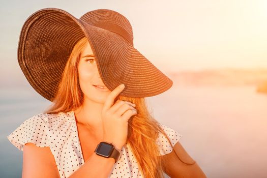 Portrait of happy young woman wearing summer black hat with large brim at beach on sunset. Closeup face of attractive girl with black straw hat. Happy young woman smiling and looking at camera at sea