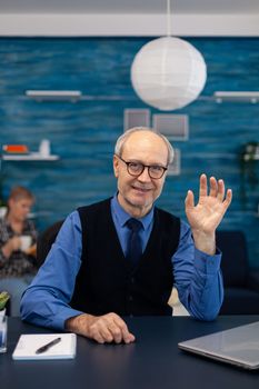 Smart senior businessman working on laptop wearing a tie and glasses. Elderly man entrepreneur in home workplace using portable computer sitting at desk while wife is holding tv remote.