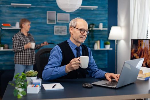 Happy senior man after reading a email with good news sitting at desk. Elderly man entrepreneur in home workplace using portable computer sitting at desk while wife is holding tv remote.