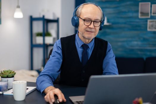 Senior businessman listening music wearing headphones working on laptop. Elderly man entrepreneur in home workplace using portable computer sitting at desk while wife is reading a book sitting on sofa.