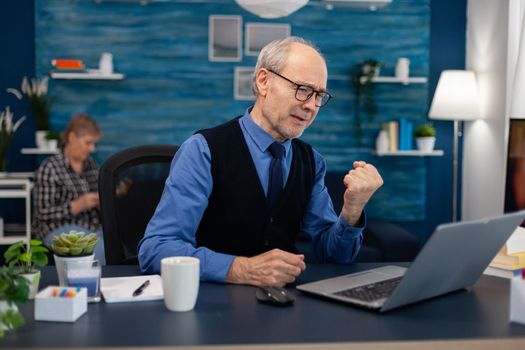 Excited senior man celebrating good news while working on laptop from home office. Elderly man entrepreneur in home workplace using portable computer sitting at desk while wife is reading a book sitting on sofa.