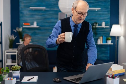 Retired businessman turning on laptop enjoying a cup of coffee. Elderly man entrepreneur in home workplace using portable computer sitting at desk while wife is holding tv remote.