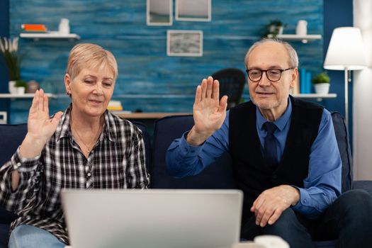 Cheerful senior couple in living room waving at webcam during online call. Happy elderly man and woman saying hello to laptop webcam sitting on sofa in the course of video conference.