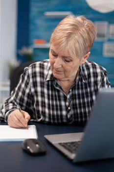 Mature entrepreneur taking notes on notebook working in home office. Elderly woman in home living room using moder technoloy laptop for communication sitting at desk indoors.