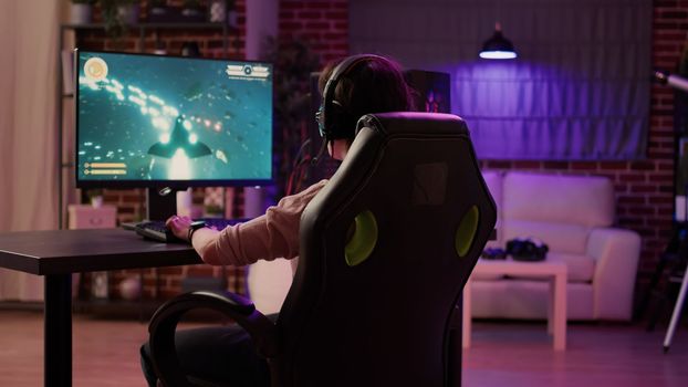 Over shoulder view of gamer girl relaxing playing rpg action game on professional pc setup while talking using headset. Woman streaming on internet fast paced space shooter simulation gameplay.