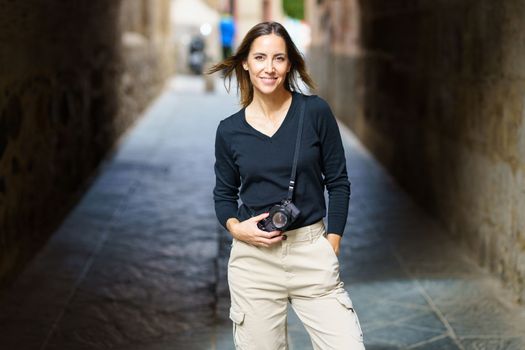 Self assured young female photographer with camera in casual clothes standing on narrow street with hand in pocket and looking at camera during vacation