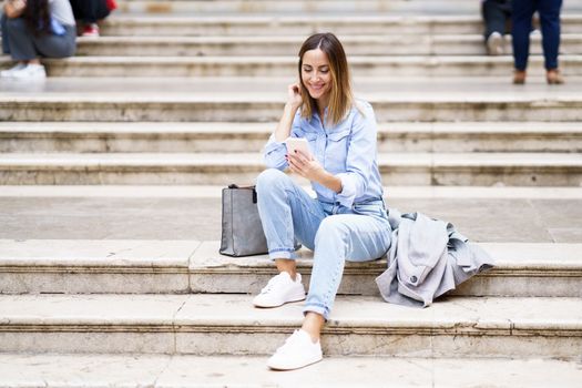 Full body glad adult woman in casual clothes touching hair and watching video on smartphone, while sitting on steps near stylish bag and coat in daytime on city street