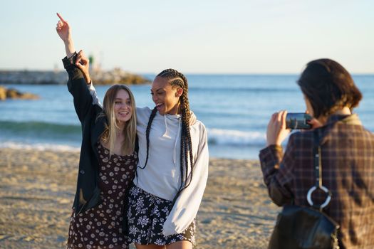 Unrecognizable woman with smartphone taking photo of smiling diverse female friends standing with raised arm on sandy coast near sea