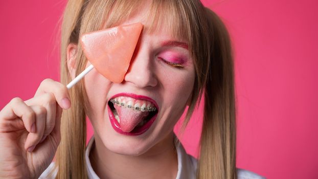 Portrait of a young woman with braces and bright makeup eating a lollipop on a pink background