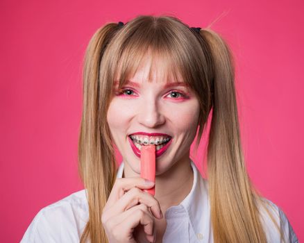 Portrait of a young woman with braces and bright makeup chewing gum on a pink background