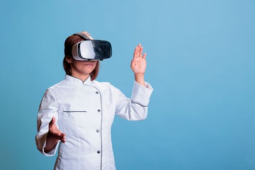 Cheerful cook woman using virtual reality headset while cooking healthy dinner in studio with blue background. Asian chef preparing culinary recipe for restaurant. Food industry concept