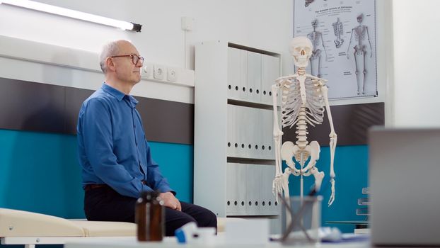 Portrait of senior patient sitting in cabinet to do consultation with medic, having appointment with specialist in doctor office. Man waiting to attend checkup visit consultation at clinic.