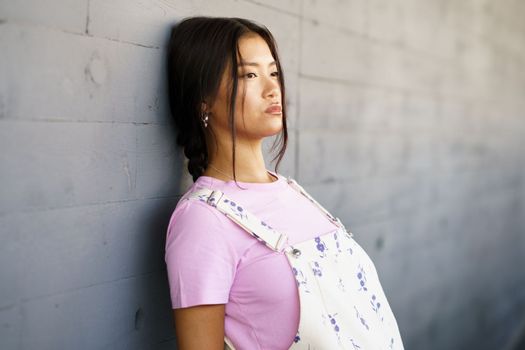 Young Chinese woman with a lost look and a serious expression leaning against the wall of a modern office building. Female wearing casual clothes.