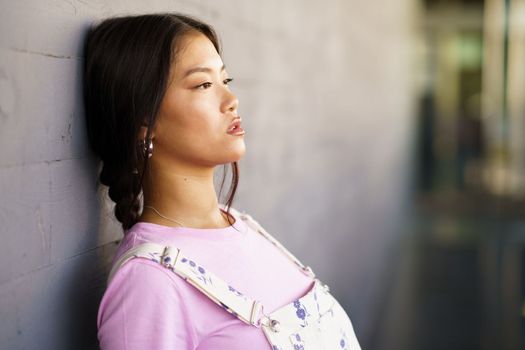 Young Chinese woman with a lost look and a serious expression leaning against the wall of a modern office building. Female wearing casual clothes.