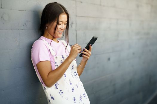 Positive Asian woman using her smartphone with a pen or stylus, outdoors. Chinese female wearing casual clothes.