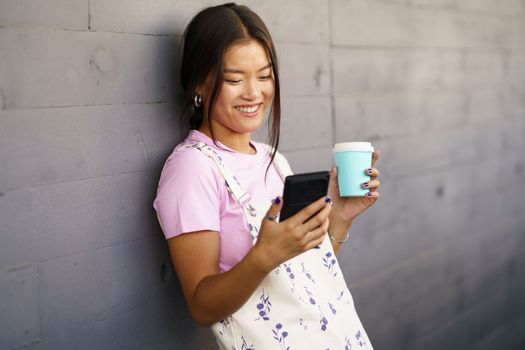 Optimistic young Asian female in stylish clothes with coffee to go smiling and watching video on cellphone while leaning on gray wall on city street
