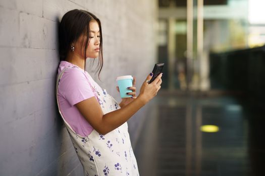 Asian female student taking a coffee break while consulting her smartphone. Chinese woman using a paper takeaway cup.