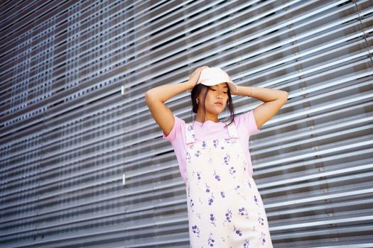 From below young Asian female in stylish clothes and cap touching head and looking down while standing near metal wall of modern building