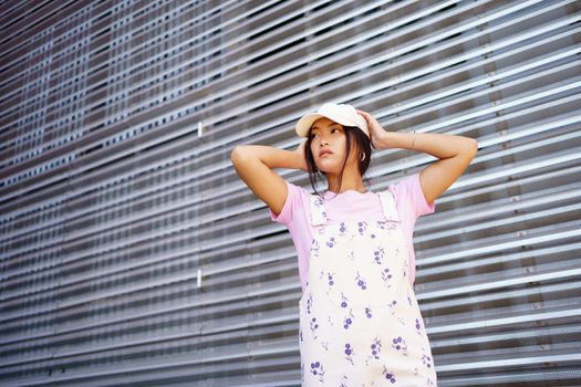 Low angle of glad Asian female in casual clothes, wearing cap, looking away standing near modern building.