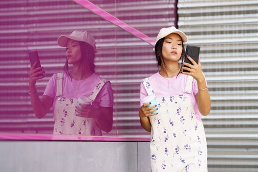 Young Asian female in stylish outfit with takeaway cup of coffee reading message on mobile phone while standing near pink glass wall in city