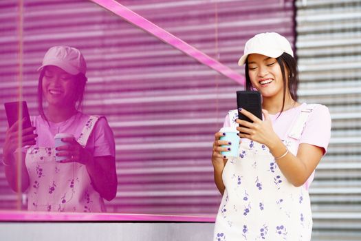 Asian female walking down the street, near a modern pink building, drinking coffee in a take-away cup, while checking her smartphone. Young Chinese woman wearing modern casual clothes.