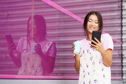 Asian girl near a modern pink building, drinking coffee in a take-away cup, while checking her smartphone. Young Chinese woman wearing modern casual clothes.