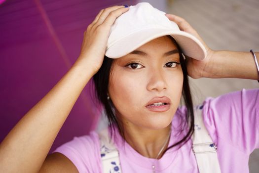 Chinese girl taking a photo or video selfie with her smartphone, to post on social media. Young Asian woman wearing modern clothes and cap.