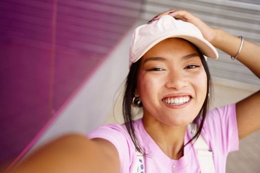 From above optimistic young Asian female in stylish cap touching head and looking at camera with smile while taking selfie near modern building on street