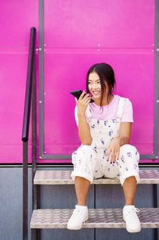 Full body young Asian female in stylish clothes, looking away with smile and recording audio message on cellphone while sitting on metal steps against pink wall