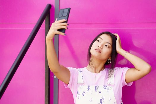 Young Asian female in stylish clothes touching head and taking selfie against pink wall of modern building