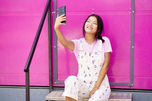 Young Chinese woman taking a photo or video selfie with her smartphone, to post on social media. Asian female wearing modern clothes in urban background.