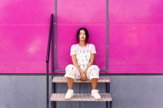 Full body stylish Asian female with dark hair sitting on metal stairs against pink wall and looking at camera on street