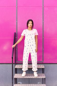 Full body young Asian woman in stylish clothes touching railing and looking at camera while standing on steps against pink wall