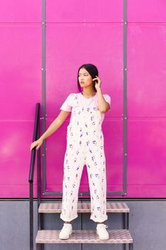 Young Chinese woman with blank stare and serious expression standing against pink wall of modern building. Asian Female in casual clothes.