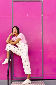 Full body young Asian female in stylish clothes holding leg on banister and leaning on hand while standing on metal step against pink wall