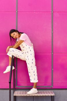 Full body happy young Asian woman, in stylish clothes holding leg on railing and looking at camera with smile while standing on metal step against bright pink wall