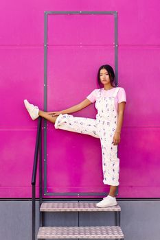 Fashion portrait of young Asian woman looking at camera with serious expression, raising one leg over railing near pink wall. Woman in casual clothes.