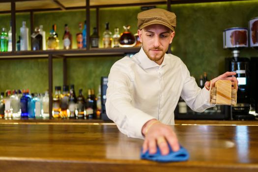Bearded man in white shirt and cap removing napkin holder and wiping counter with cloth while working in bar in evening
