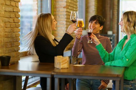 Optimistic women in casual clothes clinking glasses of alcohol drinks and proposing toast while standing near table and window on weekend day in pub
