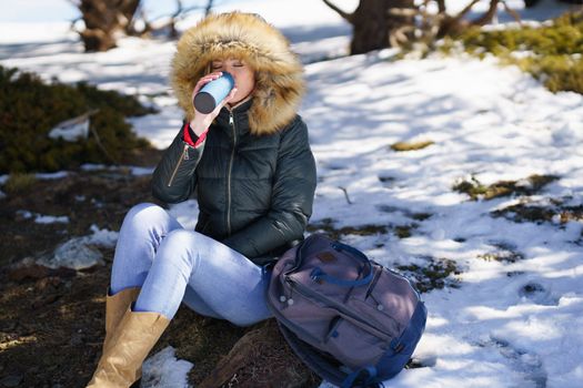 Young woman drinking something hot from a metal thermos bottle sitting on a rock in the snowy mountains., in Sierra Nevada, Granada, Spain. Female wearing winter clothes.