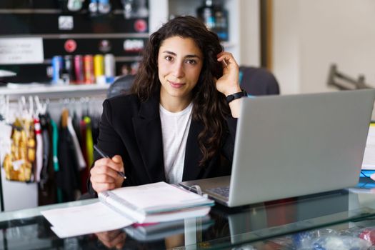Positive brunette looking at camera and adjusting long wavy hair while sitting at desk with netbook and making notes during psychology session in gym